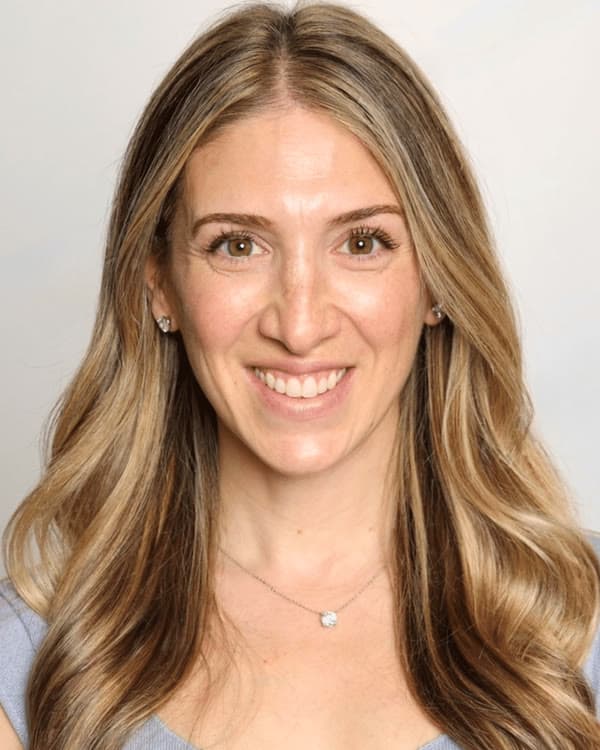 A woman with long, wavy blonde hair wearing a light purple short-sleeved top smiles in front of a plain, light background.