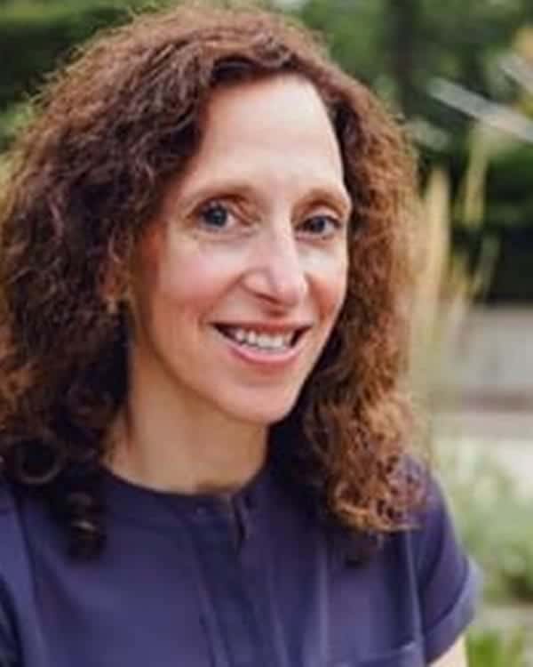 A woman with curly brown hair, wearing a short-sleeved dark top, is smiling at the camera. The background shows greenery and is slightly blurred.