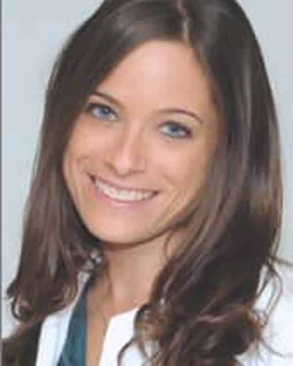 A woman with long brown hair and blue eyes is smiling at the camera. She is wearing a white coat over a teal top and is posed against a plain light gray background.