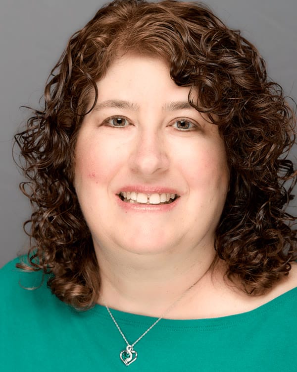 A woman with shoulder-length curly brown hair is smiling at the camera. She is wearing a green top and a silver necklace with a heart-shaped pendant, in front of a plain gray background.