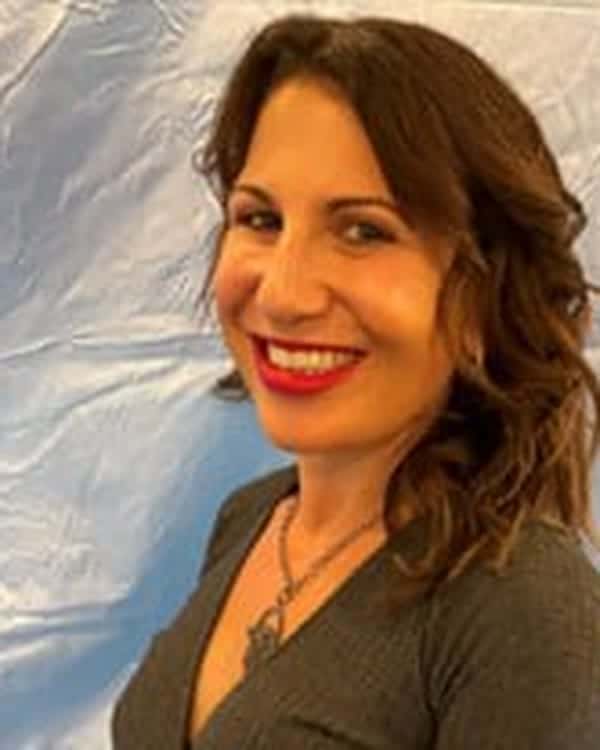 A woman with wavy brown hair is standing in front of a light blue, slightly wrinkled backdrop. She is wearing a dark long-sleeved V-neck top and smiling at the camera.