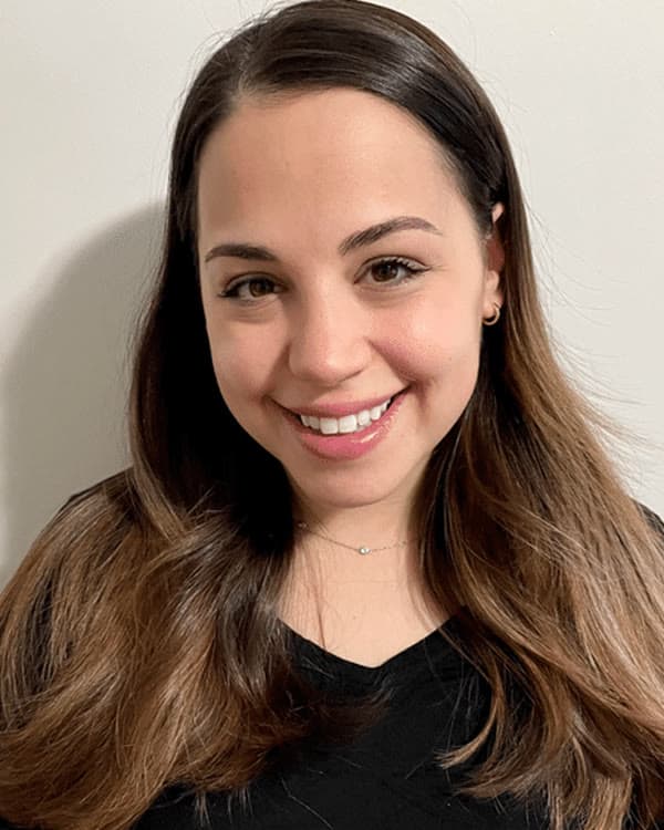 A woman with long, straight brown hair and a black top is standing in front of a plain light-colored wall, smiling at the camera.