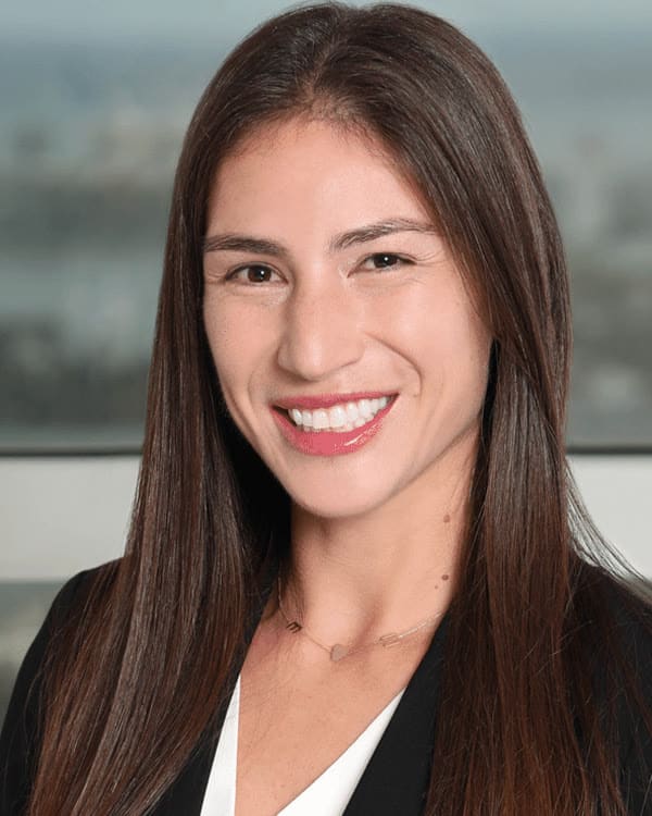 A woman with long brown hair, wearing a black blazer and white blouse, stands smiling in front of a large window with a cityscape in the background.