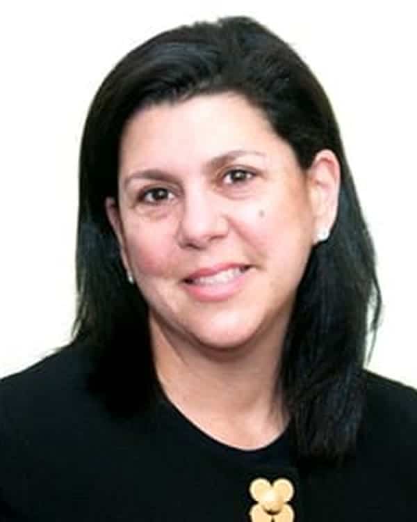 A woman with straight, shoulder-length black hair is smiling slightly. She is wearing a black top with a decorative gold detail near the neckline. The background is plain and light-colored.