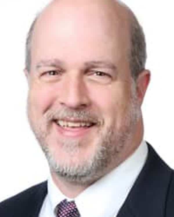 A middle-aged man with a bald head and gray beard, wearing a dark suit, white shirt, and patterned tie, smiling at the camera against a white background.