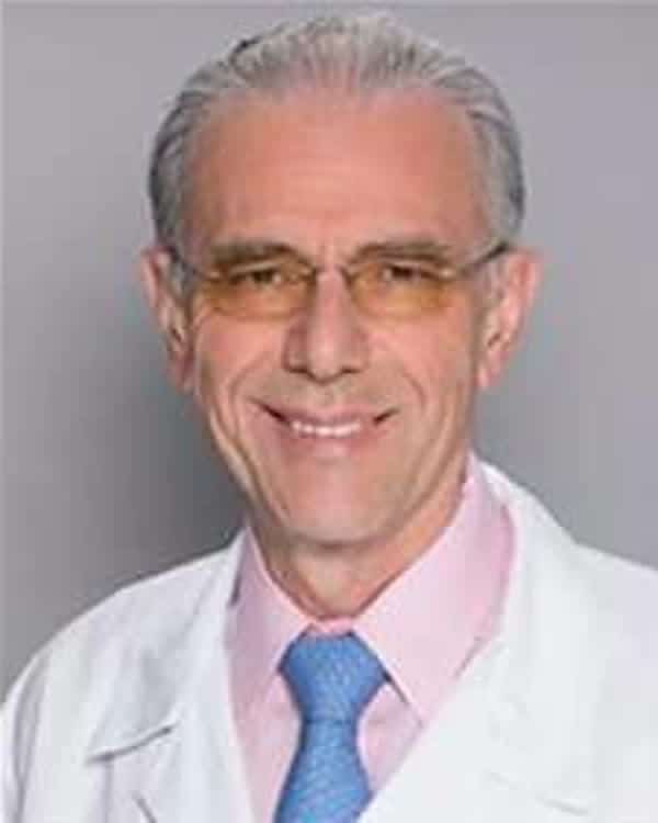 An older man with gray hair and glasses is wearing a white lab coat, a light pink shirt, and a blue tie. He is smiling and posing in front of a plain gray background.
