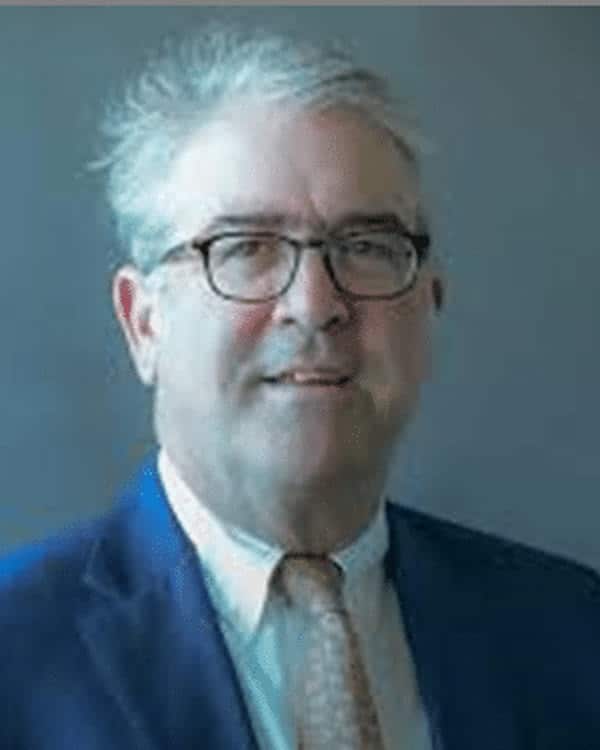 A middle-aged man with gray hair and glasses wearing a blue suit, white shirt, and patterned tie poses in front of a plain gray background.