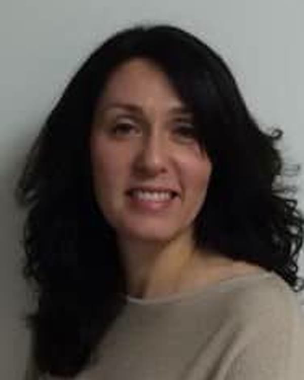 A woman with long, wavy dark hair is wearing a light beige top and is smiling softly at the camera. She is standing in front of a plain, light-colored background.