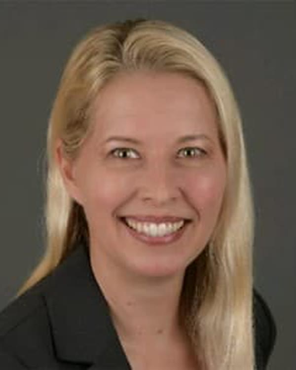 A woman with long, straight blonde hair and light skin smiles at the camera. She is wearing a dark blazer and has a neutral gray background behind her.