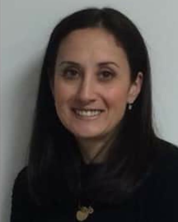 A woman with long, straight dark hair and light skin is smiling at the camera. She is wearing a black top, small earrings, and a necklace with two round pendants. The background is plain and gray.