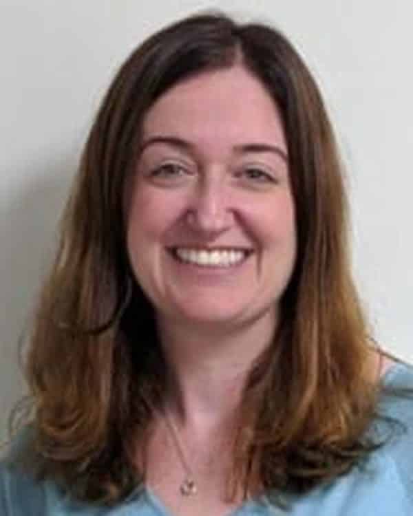 A woman with long brown hair wearing a light blue shirt is smiling in front of a plain light-colored wall.