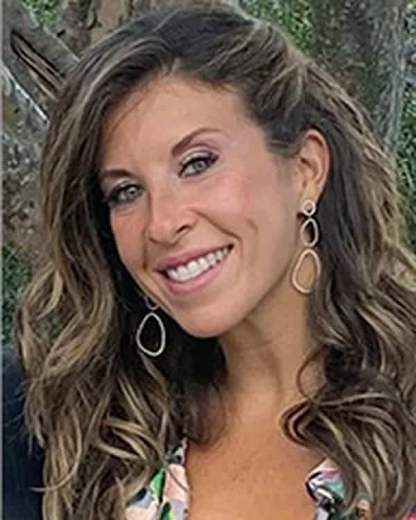 A woman with long wavy brown hair and dangling earrings smiles at the camera. She is outdoors, standing in front of trees, and is wearing a patterned top with a deep neckline.