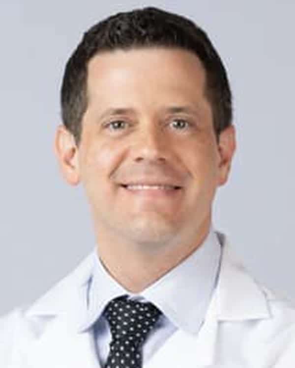 A man with short dark hair wearing a white lab coat, light blue dress shirt, and dark polka dot tie, smiling and facing the camera against a plain light background.
