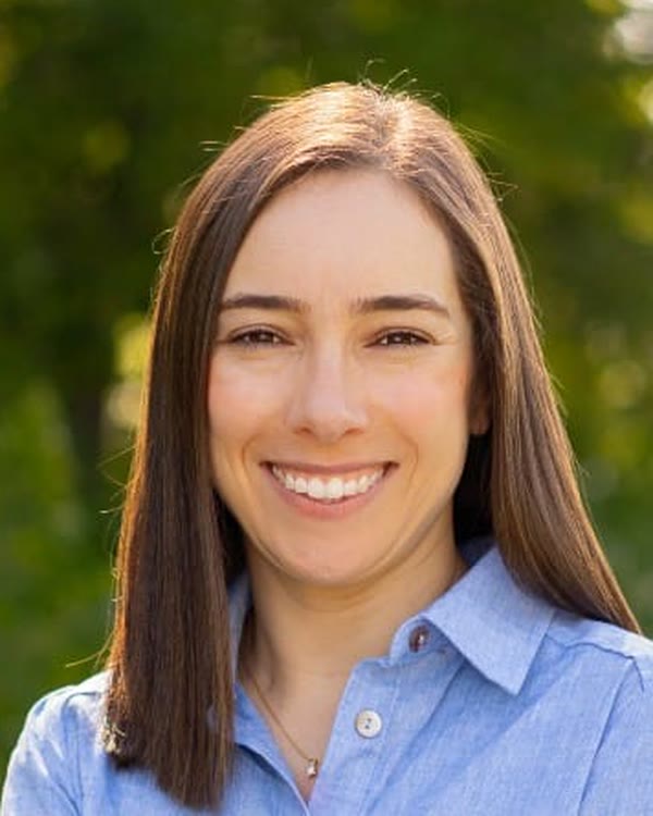 A person with long brown hair and wearing a blue shirt. standing outside in front of greenery.