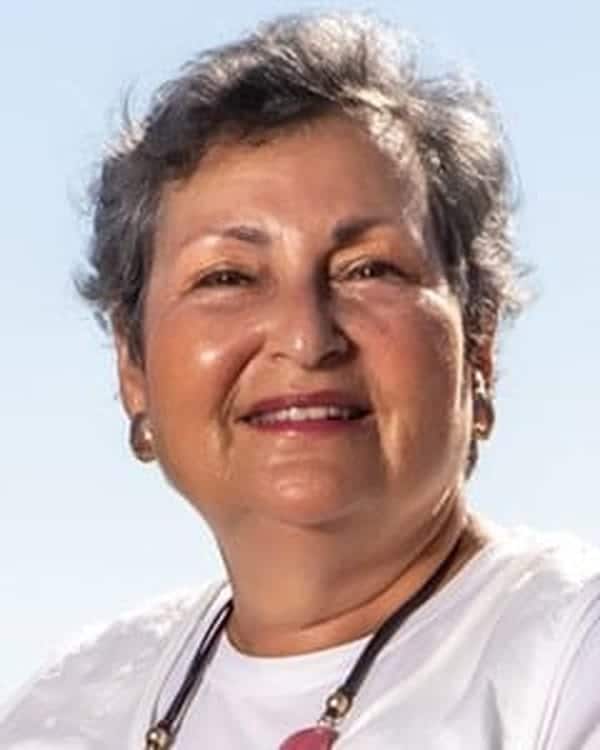 An older woman with short gray hair smiles at the camera. She is wearing a white top, a necklace, and gold hoop earrings. The background is a clear blue sky.