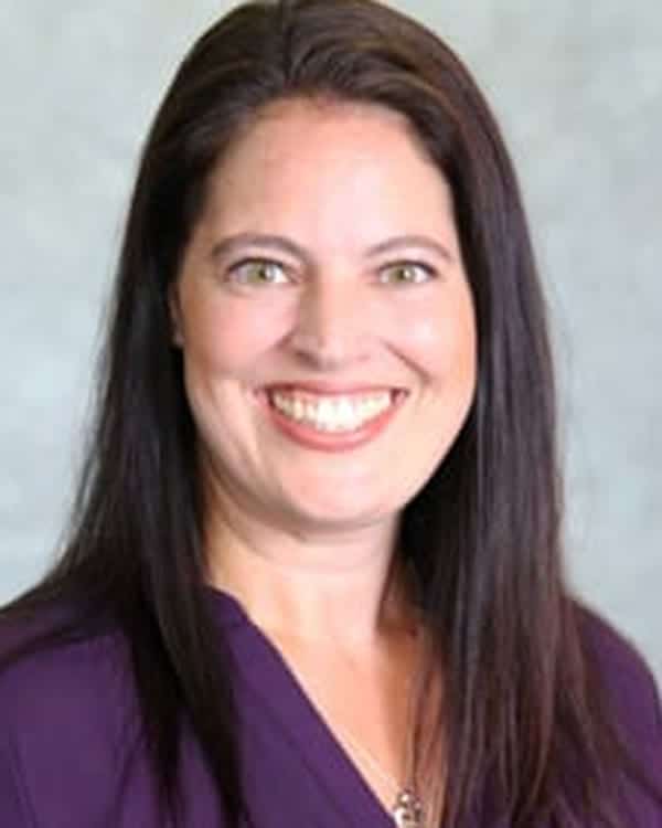 A woman with long dark brown hair and light skin is smiling at the camera. She is wearing a purple blouse and is posed in front of a plain light gray background.