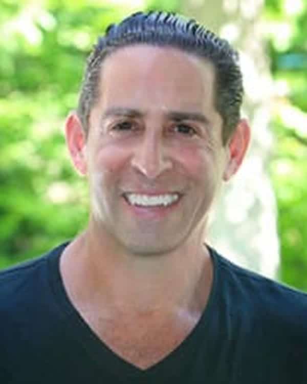 A man with dark, slicked-back hair and a black V-neck shirt is smiling at the camera. The background is outdoors with green foliage in soft focus.