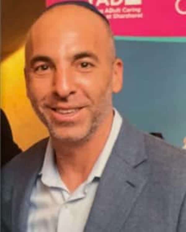 A man with short hair and a trimmed beard, wearing a light blue shirt and gray blazer, stands indoors in front of a colorful sign with partially visible text.