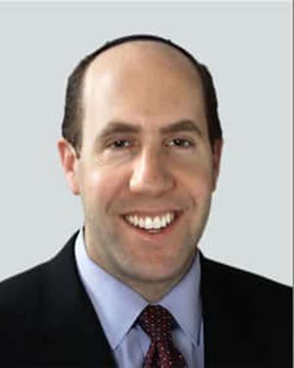 A man with short dark hair and a receding hairline is smiling. He is wearing a dark suit, light blue collared shirt, and a patterned tie. The background is plain light gray.