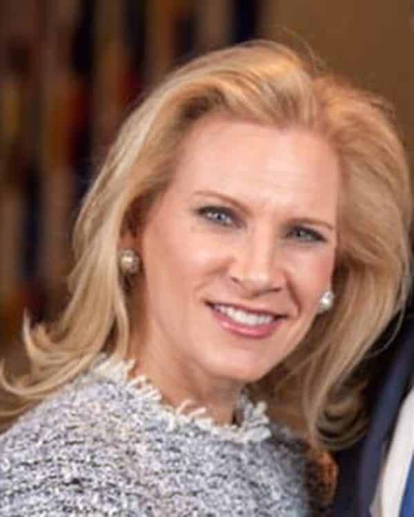 A woman with long blonde hair and light skin is smiling at the camera. She is wearing pearl earrings and a textured light-colored top. The background is blurred with muted colors.
