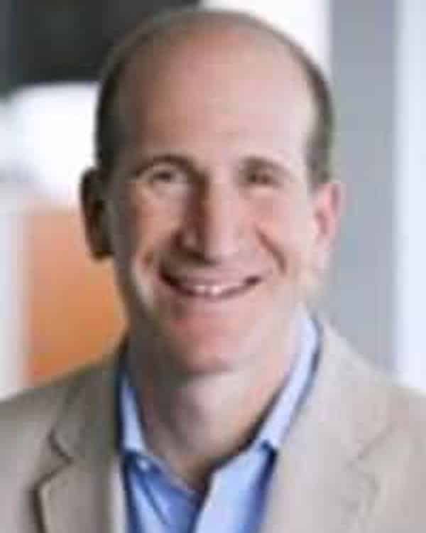 A middle-aged man with short, light brown hair is smiling. He is wearing a beige blazer over a light blue shirt. The background is out of focus with neutral colors.
