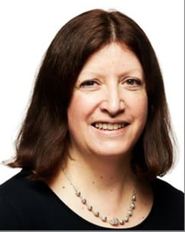 A woman with straight, shoulder-length brown hair is smiling at the camera. She is wearing a black top and a beaded necklace. The background is plain white.