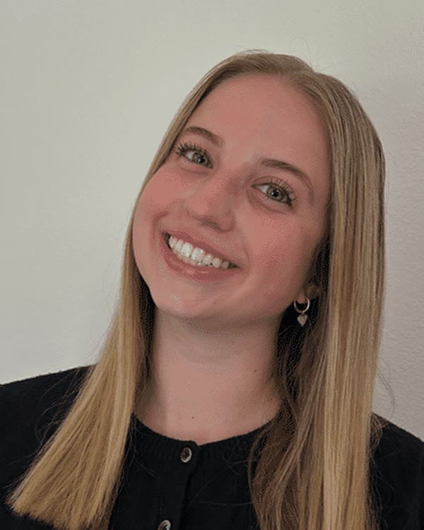 A person with straight, shoulder-length blonde hair is smiling at the camera. They are wearing a black button-up top and standing in front of a plain light-colored wall.