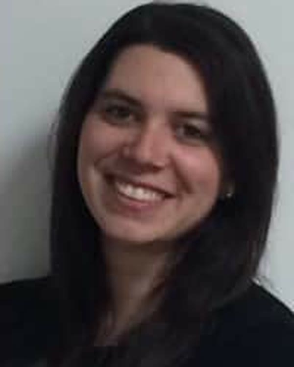 A woman with long dark hair is smiling at the camera. She is wearing a dark top and is positioned in front of a plain, light-colored background.