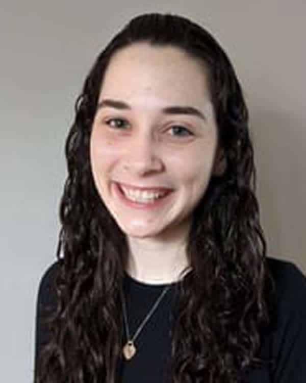 A person with long, curly dark hair wearing a black top and a necklace is standing in front of a plain, light-colored background, smiling at the camera.
