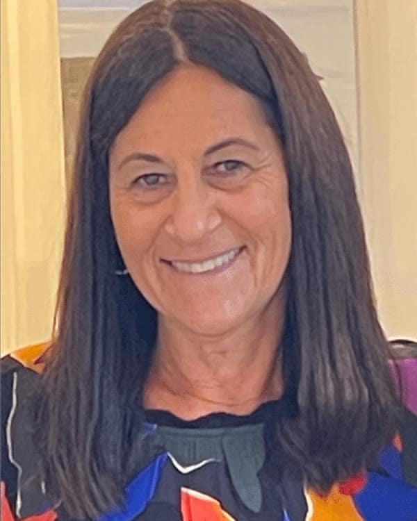 A woman with long, straight dark hair smiles at the camera. She is wearing a black top with a bold, colorful floral pattern. The background features a softly lit interior and a landscape painting on the wall.