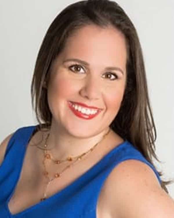 A woman with long brown hair is smiling at the camera. She is wearing a sleeveless blue top and a gold necklace. The background is plain and light gray.
