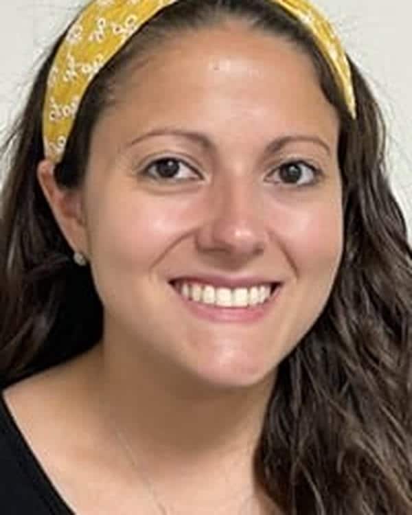 A woman with long brown hair, wearing a yellow headband and a black top, smiles in front of a plain light-colored background.
