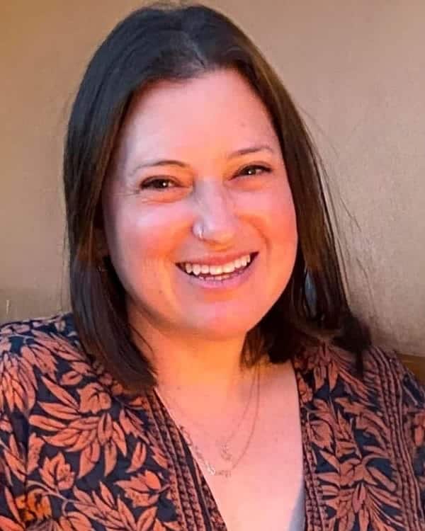 A woman with straight, shoulder-length dark hair smiles at the camera. She is wearing a patterned top with orange leaves on a dark background and a gold necklace. The background is a plain, light brown wall.