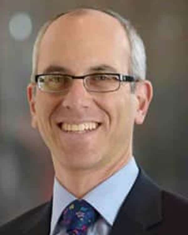 A middle-aged man with short gray hair and glasses is smiling. He is wearing a dark suit, a light blue shirt, and a patterned tie. The background is blurred.