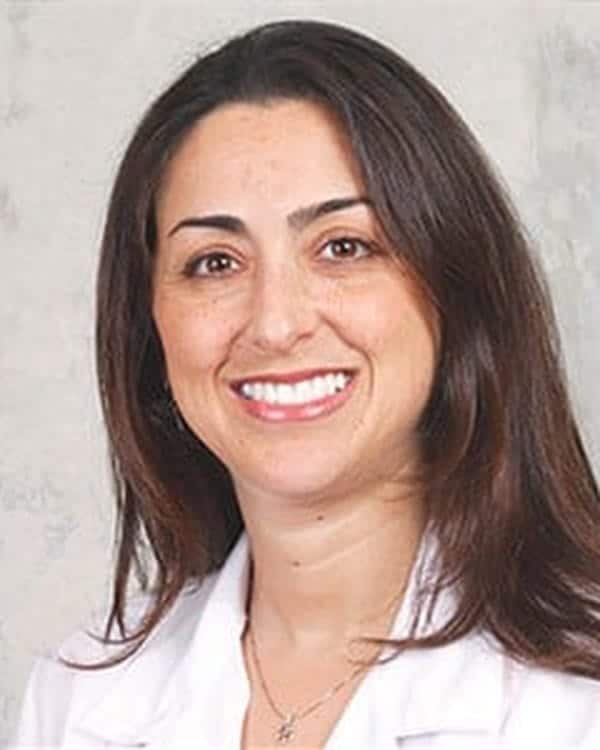 A woman with long brown hair, wearing a white lab coat, is smiling at the camera against a light gray background.