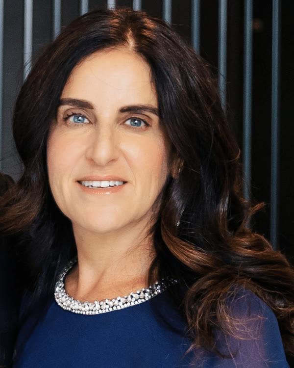 A woman with long dark wavy hair and blue eyes wears a navy blue top with a beaded neckline. She is smiling slightly and standing in front of a background with vertical black and gray lines.