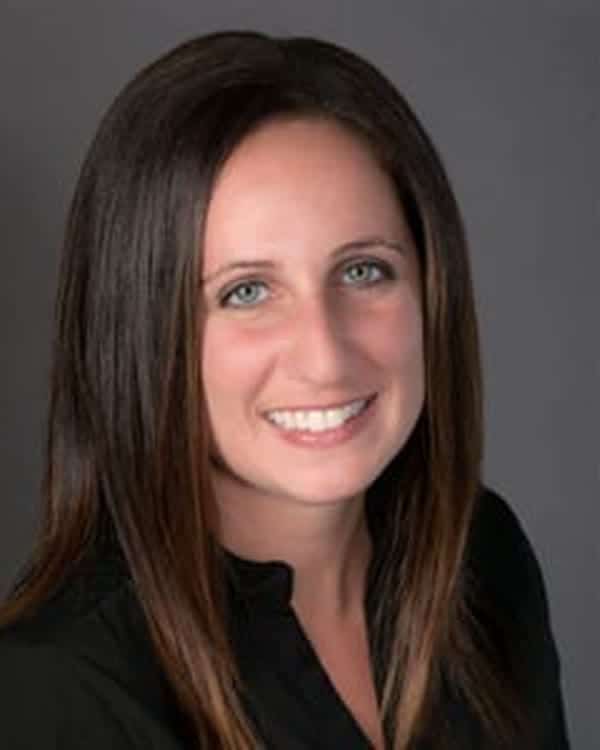 A woman with straight, dark brown hair and light skin is smiling at the camera. She is wearing a black top and is posed in front of a plain gray background.