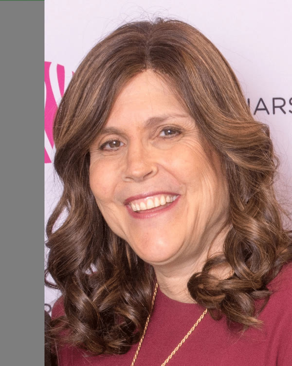A woman with long, wavy brown hair and a maroon top is smiling in front of a light-colored background with partial text and design elements visible.
